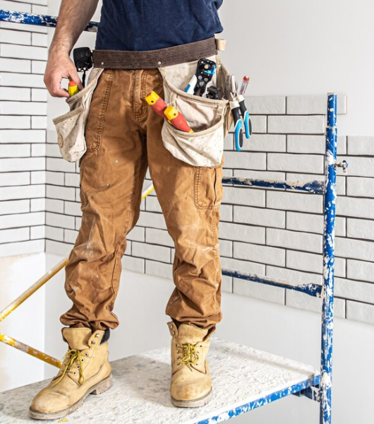 Construction worker wearing a tool belt with various hand tools while working indoors near a tiled wall